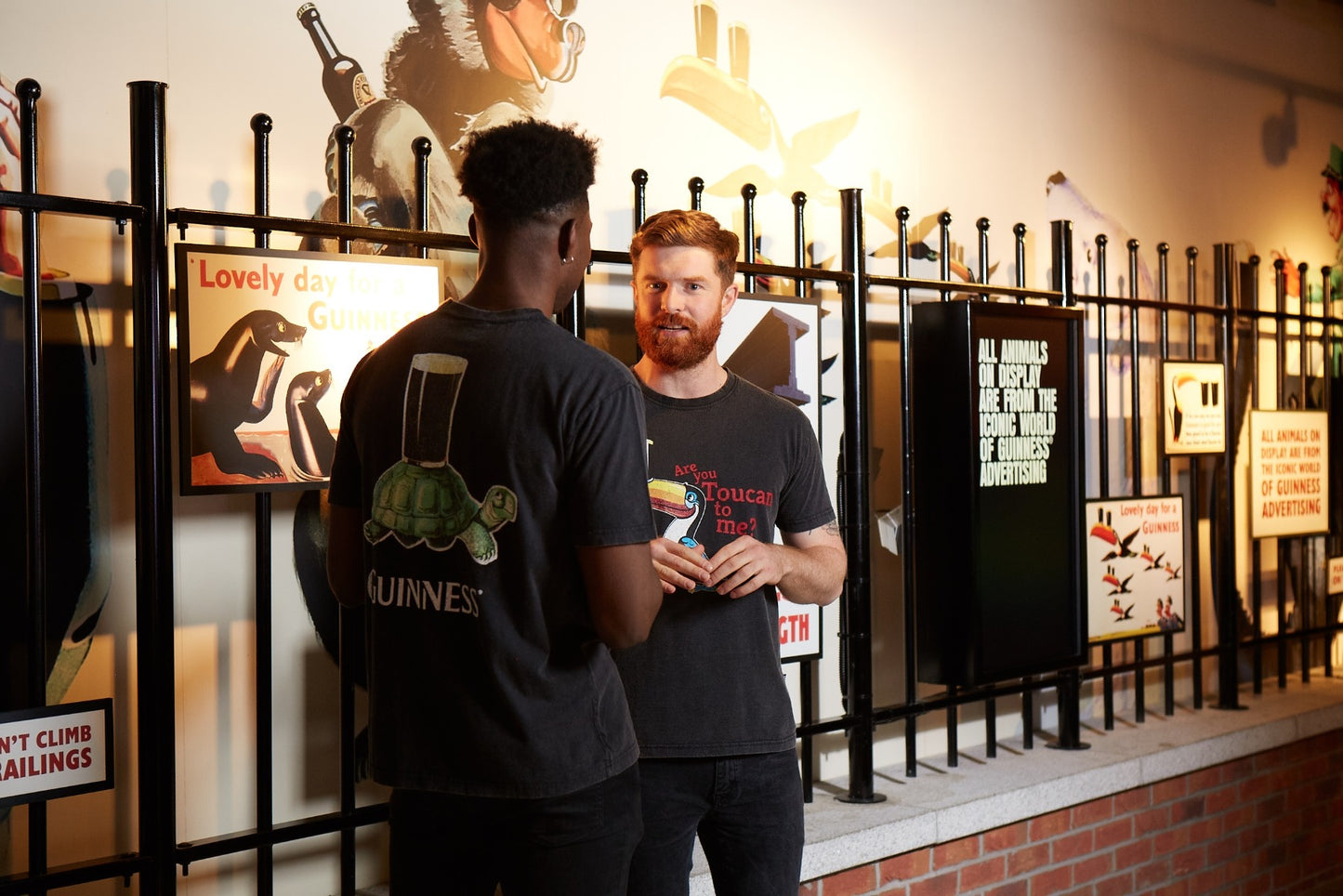 Two men stand chatting before a wall of vintage Guinness ads; one wears the Guinness Premium Vintage Tortoise Back Graphic Tee, crafted from soft 100% cotton with a subtle distressed effect. Black railings complete the scene.