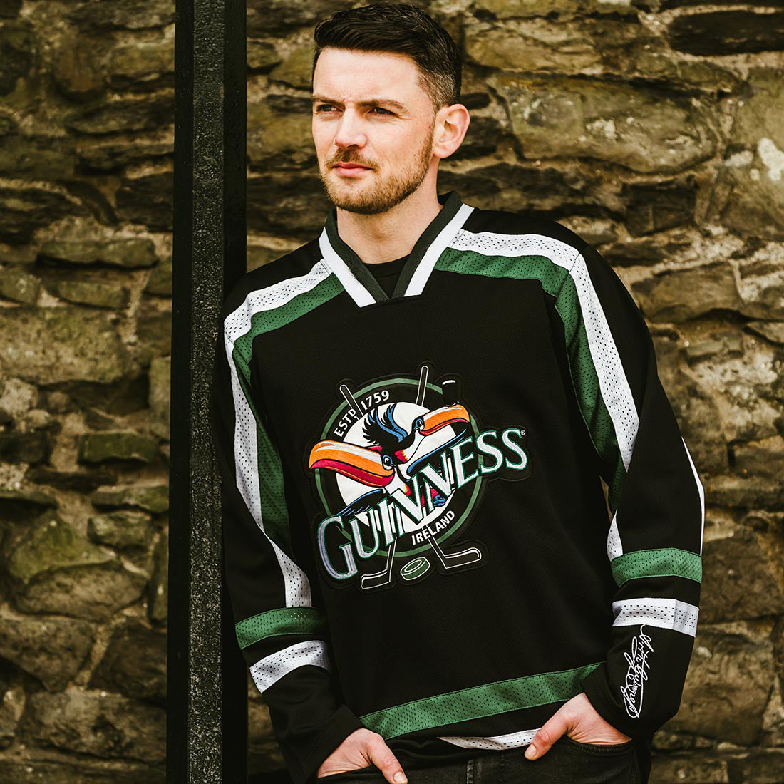 A man wearing the Guinness Toucan Hockey Jersey in black and green by Guinness stands against a stone wall, looking to the side with a serious expression.