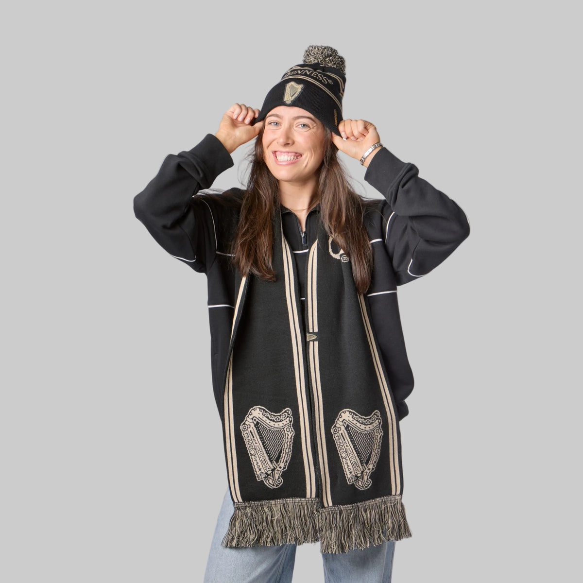 Woman smiling, wearing the Guinness Black & Gold Sports Beanie – Vintage Harp Knit, black scarf with harp designs, black sweater, and light blue jeans against a white background.