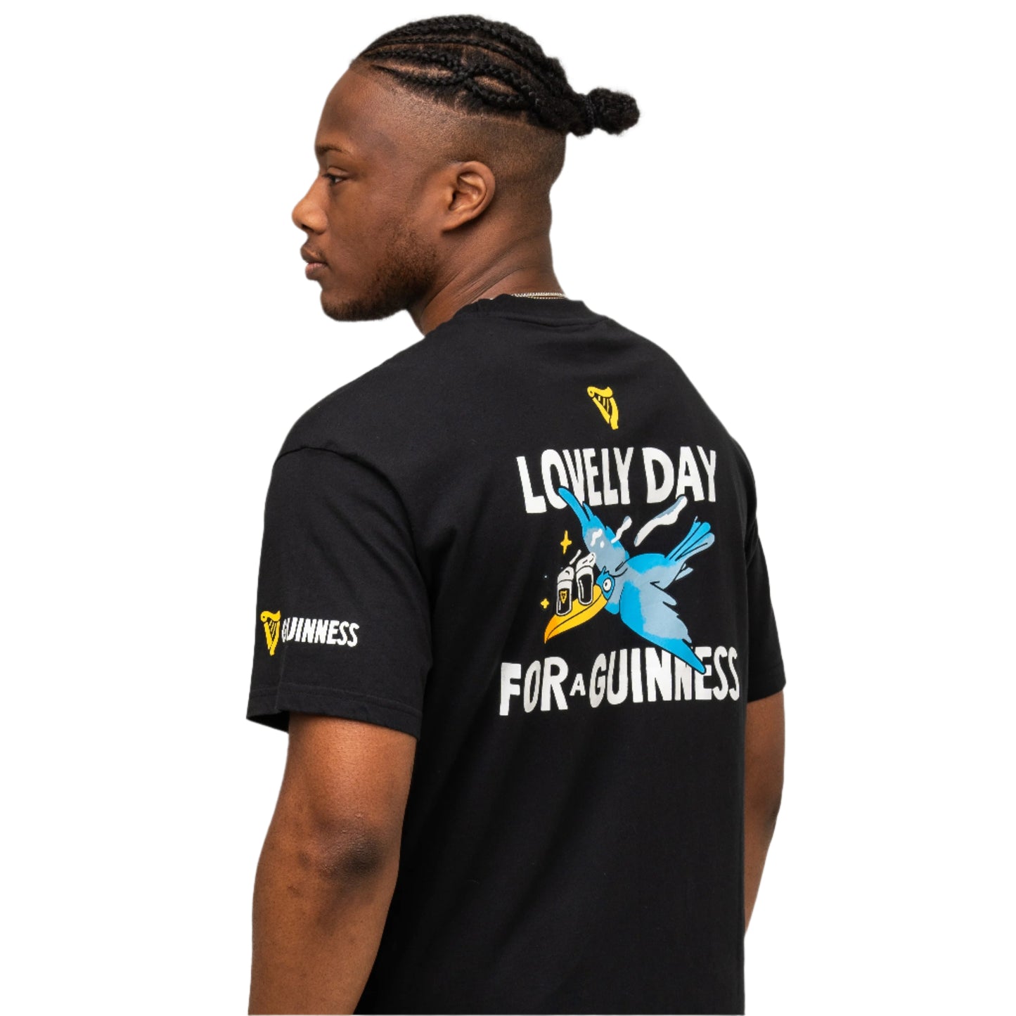 A man faces away, wearing the Guinness Seb Curi Oversize Tee - Black from Guinness Webstore US, featuring “Lovely Day for a Guinness” and a flying toucan graphic on the back, plus logos on the sleeve and upper back.