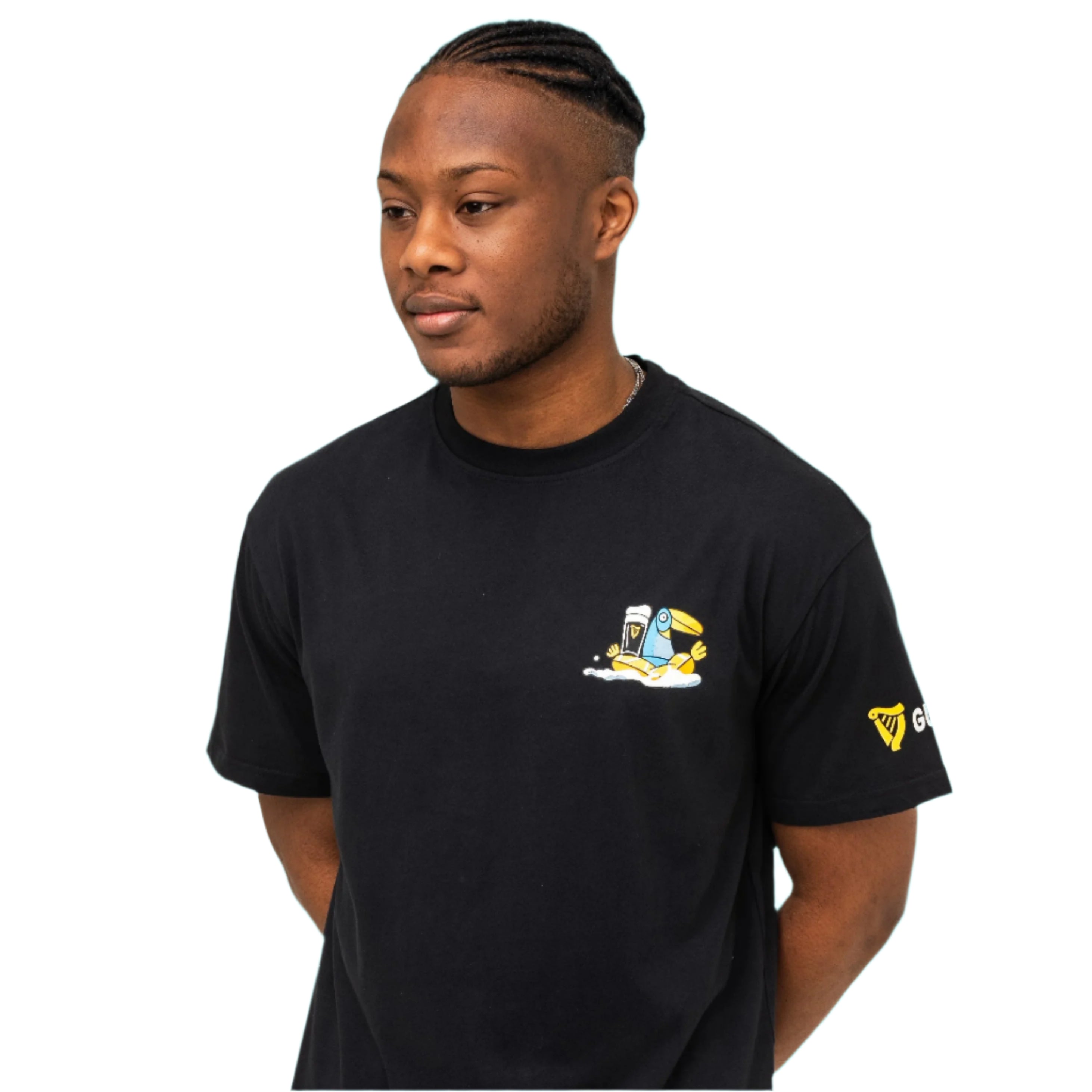 A man with short braids wears the Guinness Seb Curi Oversize Tee - Black by Guinness Webstore US, featuring a small cartoon chest graphic from the Sebastian Curi collaboration, against a plain white background.