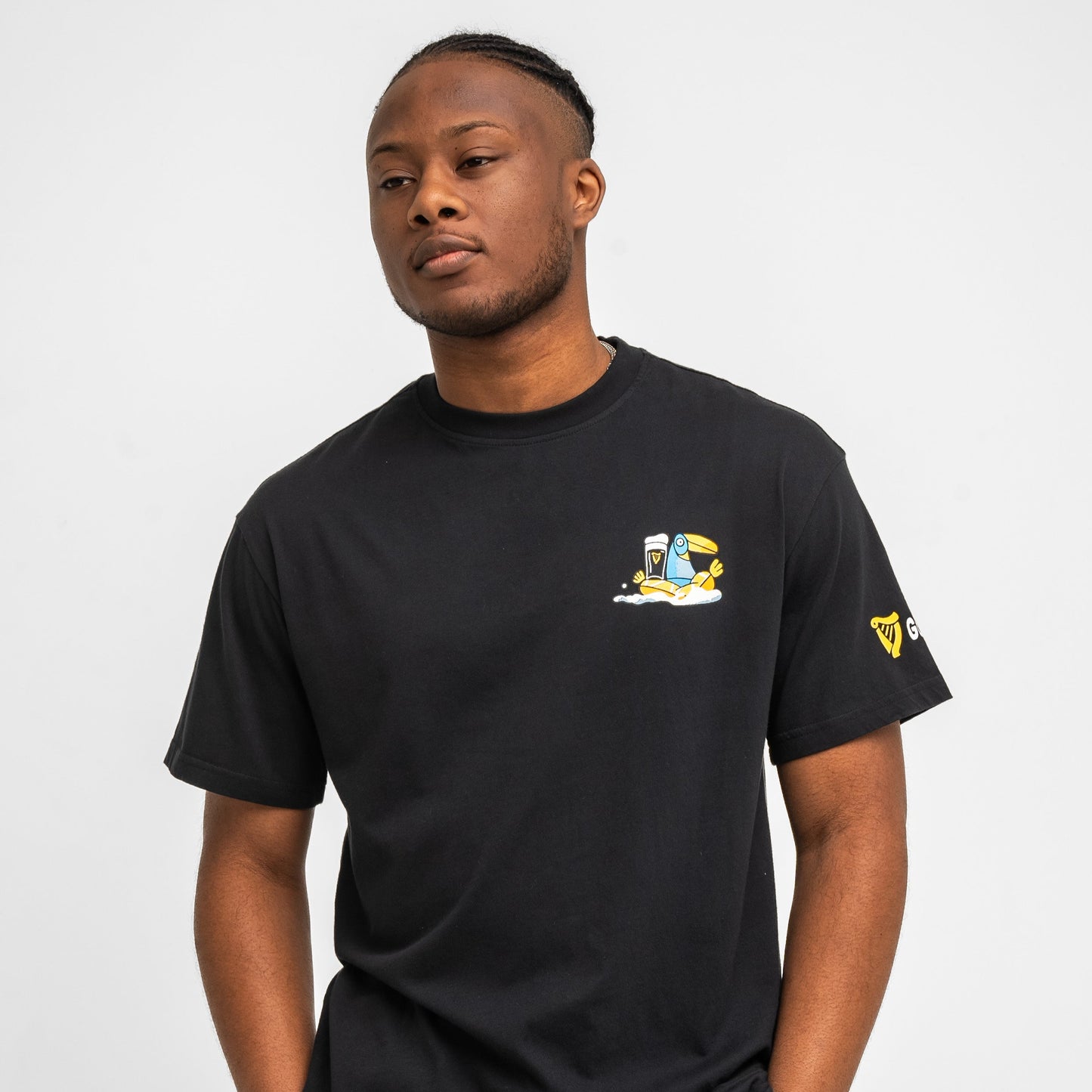 A man wearing the Guinness Seb Curi Oversize Tee - Black from Guinness Webstore US, featuring a small cartoon character and clock graphic on the left chest, stands against a plain white background. Part of the Sebastian Curi collaboration.
