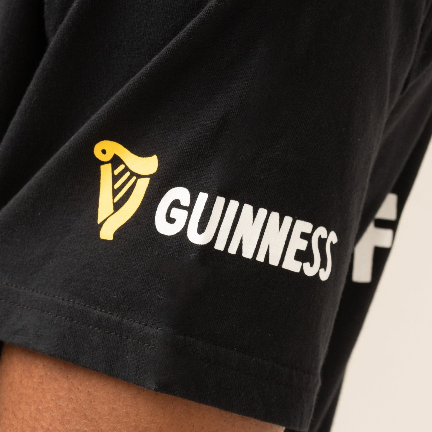 Close-up of a black Guinness Seb Curi Oversize Tee sleeve with a yellow harp logo and “GUINNESS” in white, from Guinness Webstore US and designed with Sebastian Curi.