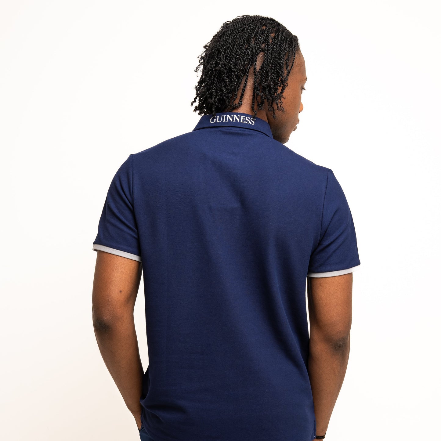 A person faces away in a navy Guinness Performance Polo from Guinness Webstore US, featuring "GUINNESS" text on the collar and made from breathable fabric, set against a plain white background.