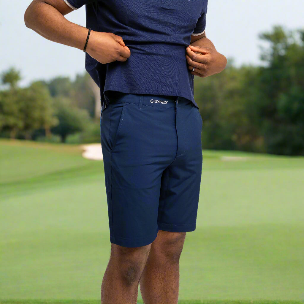 A person wearing Guinness Performance Shorts - Navy from Guinness Webstore US and a matching polo stands against a white background, lifting the shirt hem with both hands.