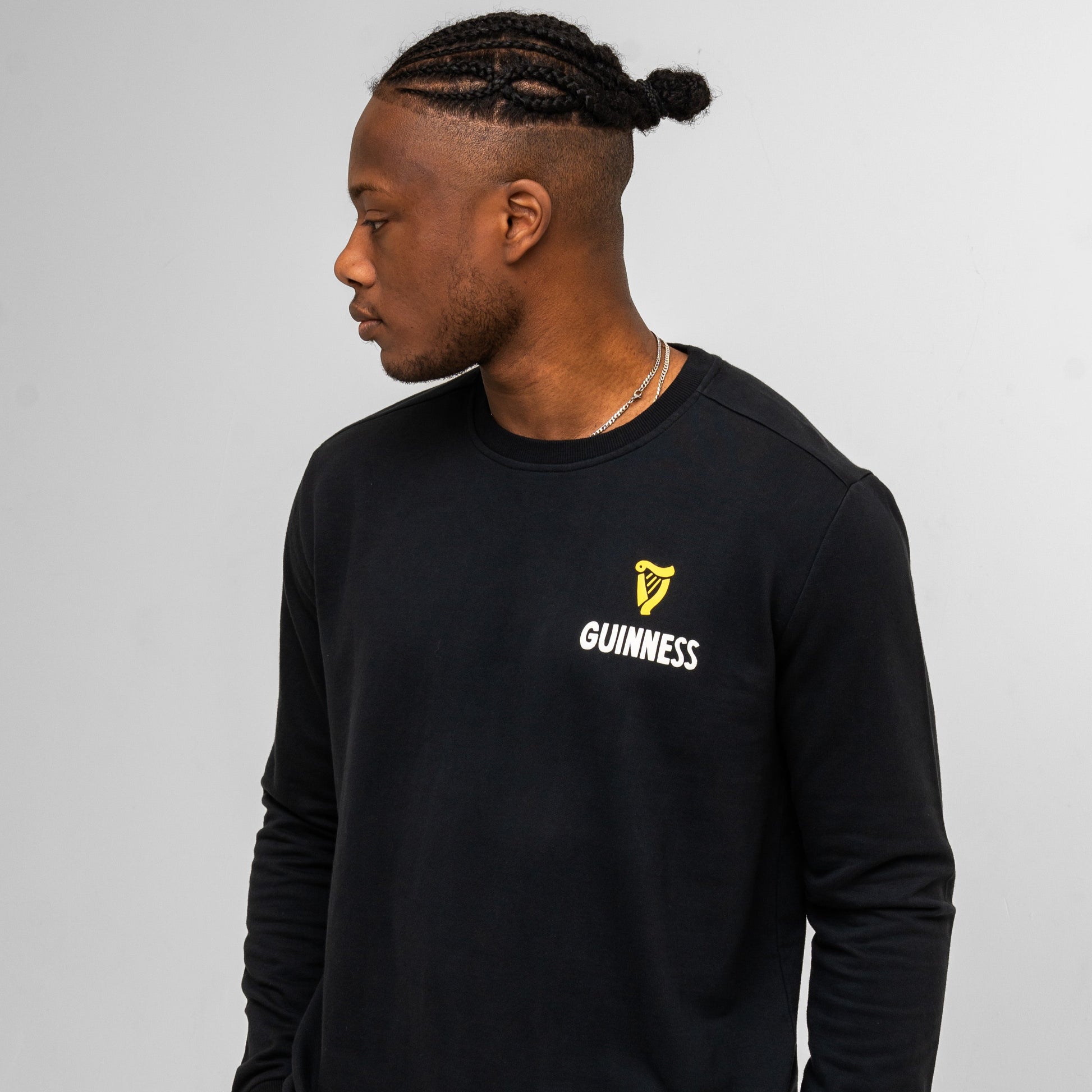 A man with braided hair, facing left, wears the Guinness Seb Curi Lovely Day Sweatshirt in black from Guinness Webstore US, featuring a yellow logo on the chest, set against a plain light grey background.