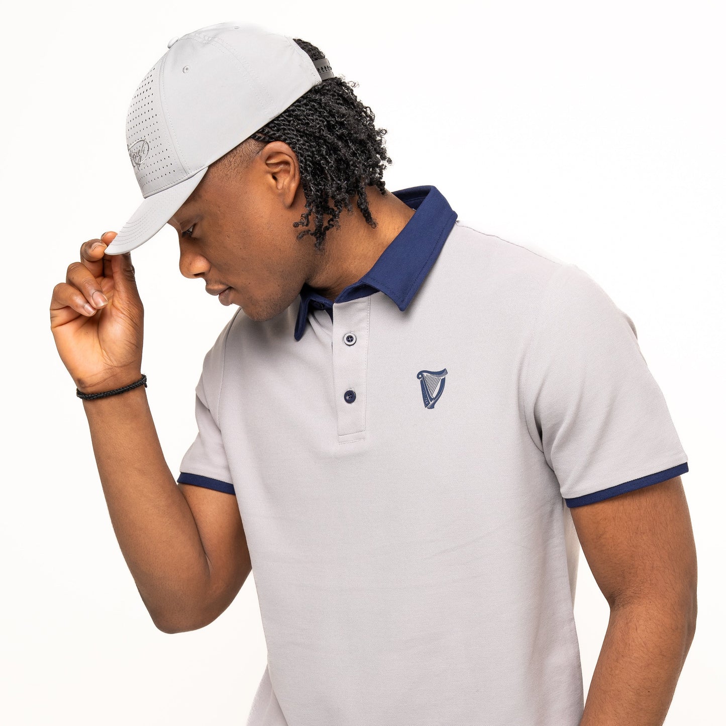 A man in the Guinness Performance Polo - Grey from Guinness Webstore US, featuring premium double pique fabric with navy trim and logo, pairs it with a matching light gray cap and navy pants, adjusting his cap.