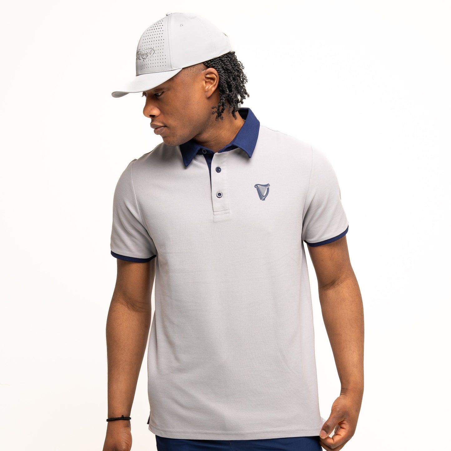A man wearing the Guinness Performance Polo - Grey by Guinness Webstore US, paired with navy blue shorts and a light gray baseball cap, stands against a plain white background.