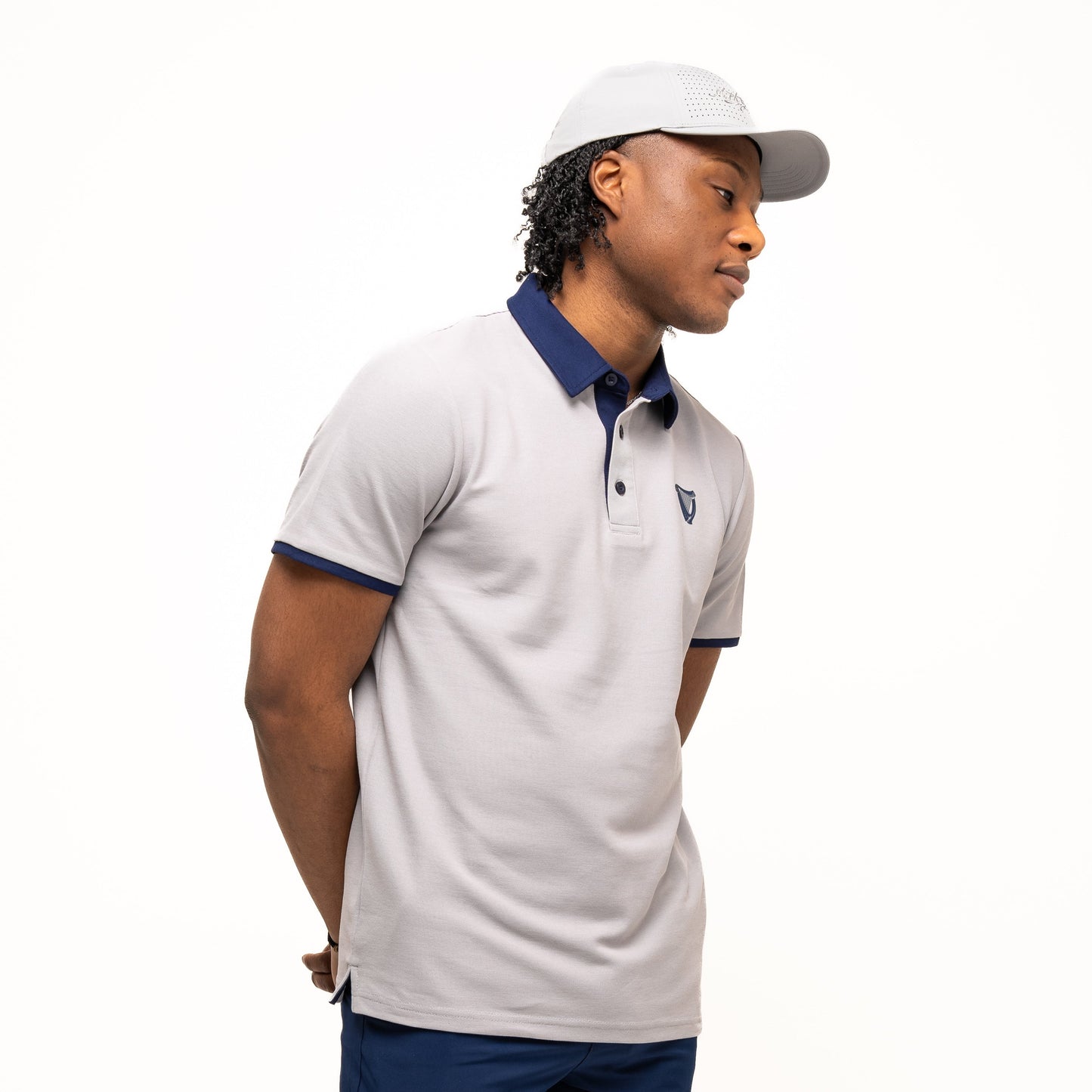 A man wearing the Guinness Performance Polo - Grey by Guinness Webstore US, along with navy blue shorts and a light gray cap, stands with his hands behind his back against a plain white background.