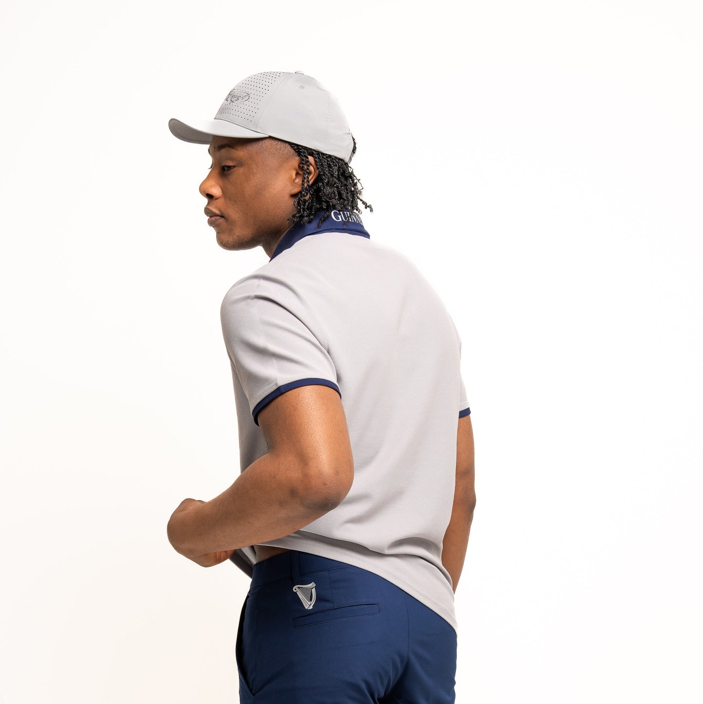 A man stands facing away, looking over his right shoulder, wearing the Guinness Performance Polo - Grey from Guinness Webstore US along with navy blue shorts and a gray cap, set against a plain white background.