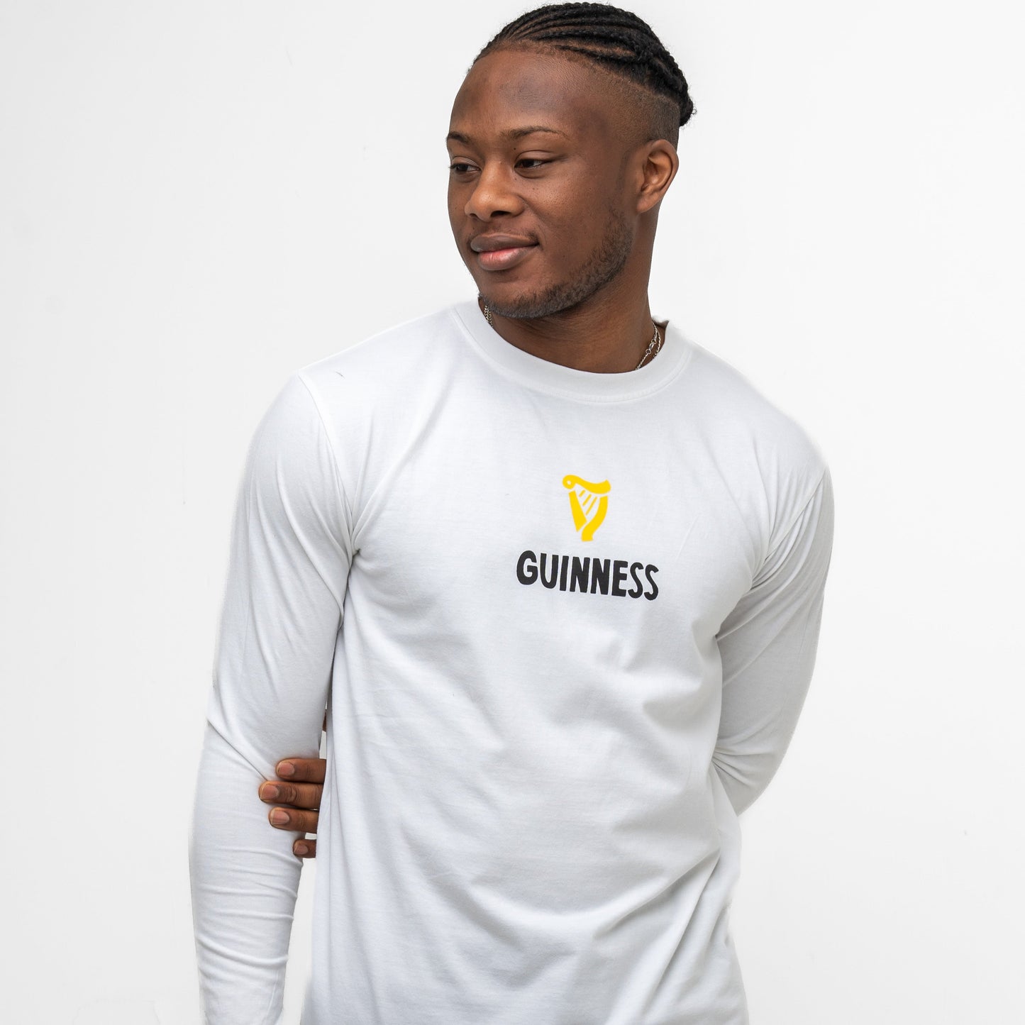 A man with braided hair models the Guinness Seb Curi Lovely Day Longsleeve Tee in white by Guinness Webstore US, featuring the yellow harp logo and black "GUINNESS" text—a wearable art piece from the Sebastian Curi collaboration.