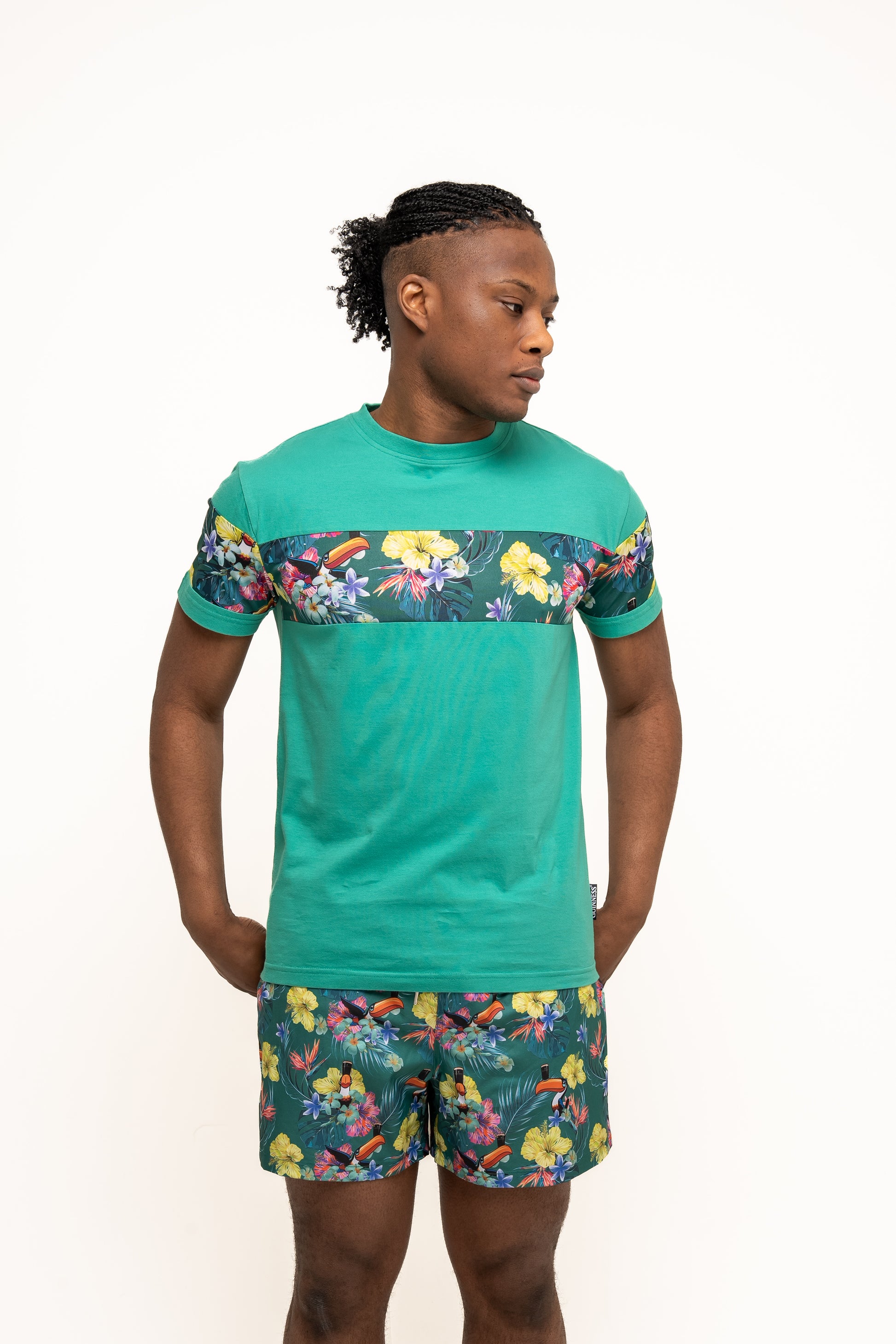 A man stands against a plain white background wearing Guinness Toucan Hawaiian Green Beach Shorts from Guinness Webstore US, paired with a green t-shirt featuring a floral pattern on the chest and sleeves.