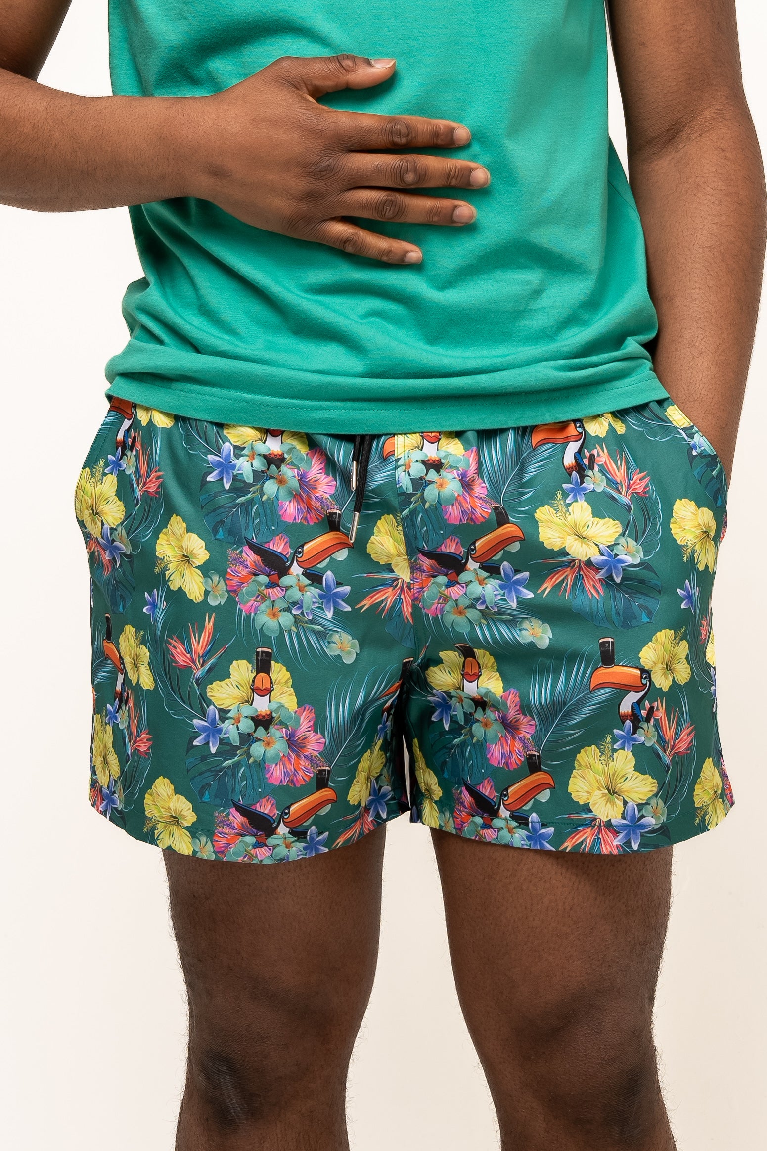 A person wearing a green shirt and Guinness Toucan Hawaiian Green Beach Shorts by Guinness Webstore US stands against a plain background, hands resting on their stomach and in their pocket.