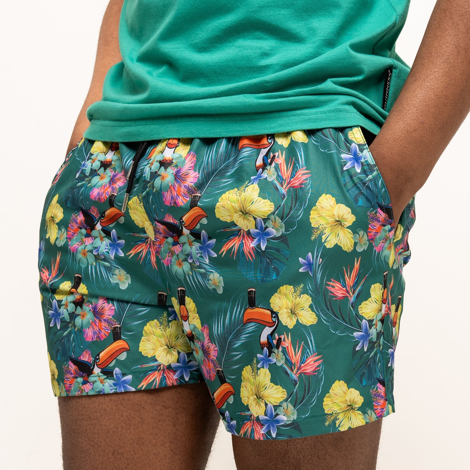 A person wearing a green t-shirt and Guinness Toucan Hawaiian Green Beach Shorts from Guinness Webstore US, hands in pockets, stands against a plain background.
