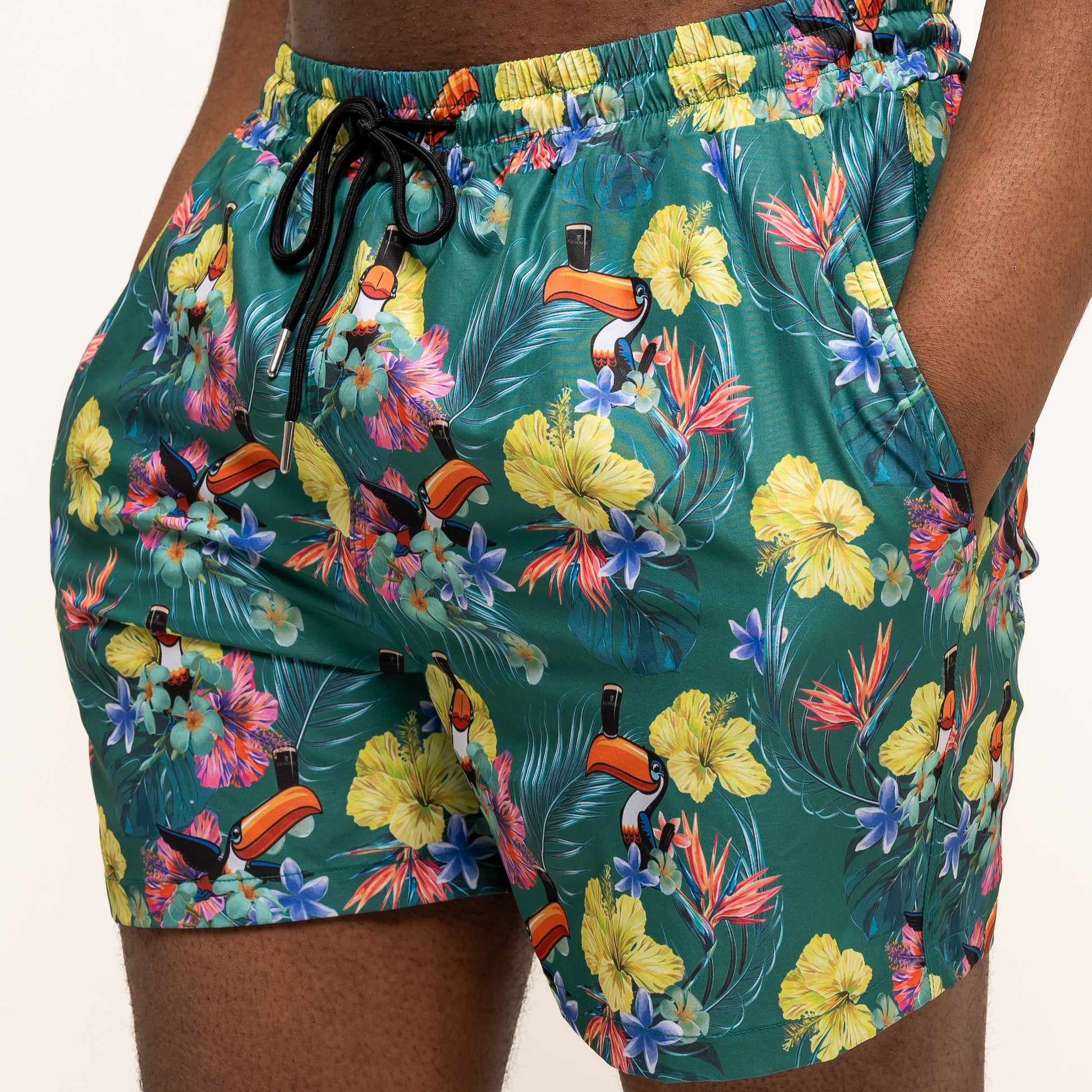 A person stands against a plain background wearing Guinness Webstore US’s Guinness Toucan Hawaiian Green Beach Shorts, featuring a vibrant tropical floral and toucan print.