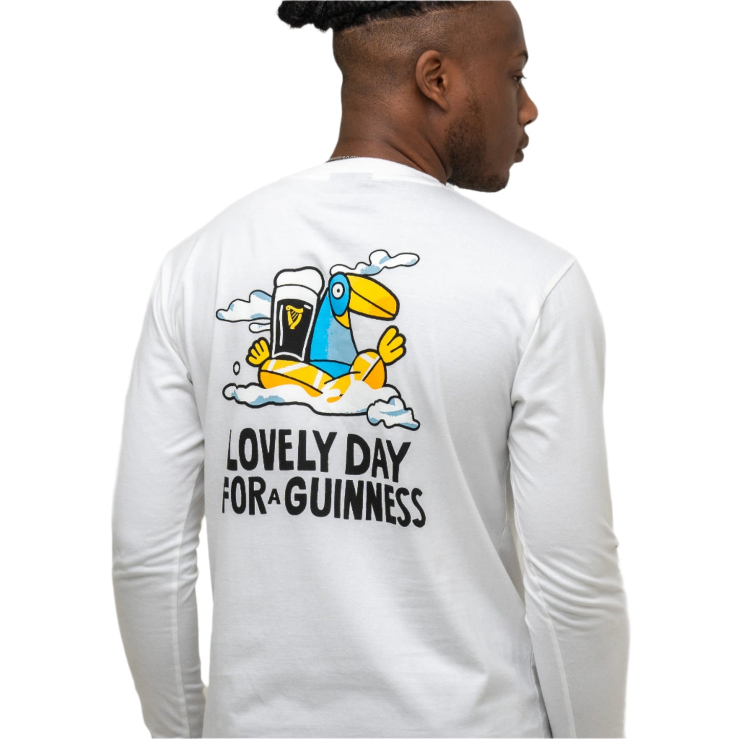 A man wears the Guinness Seb Curi Lovely Day Longsleeve Tee in white from Guinness Webstore US, featuring a cartoon toucan with a pint and "Lovely Day For a Guinness" text on the back—a unique Sebastian Curi collaboration.