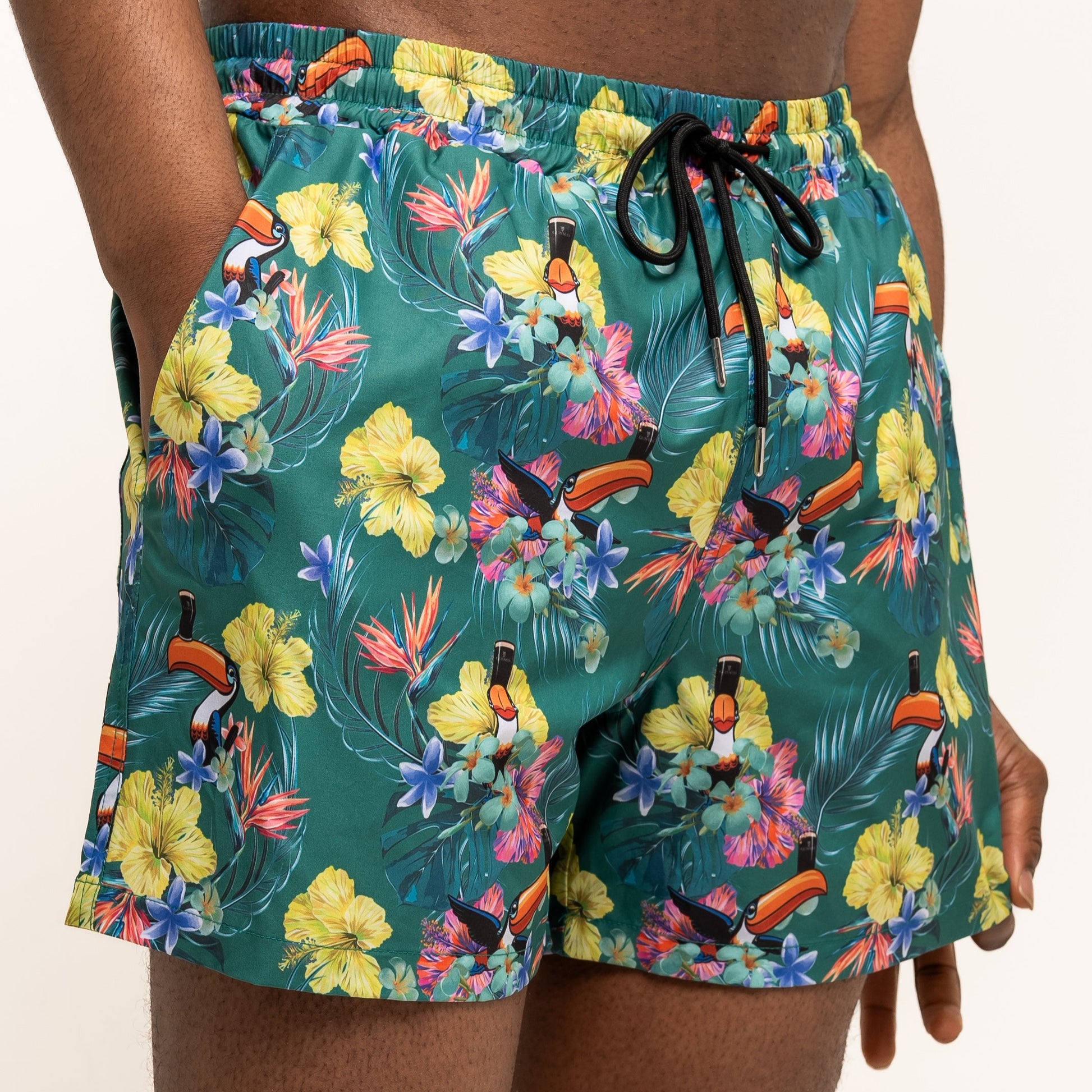 A person wearing Guinness Toucan Hawaiian Green Beach Shorts from Guinness Webstore US, featuring a vibrant tropical floral and toucan print, stands with hands in pockets against a plain background.