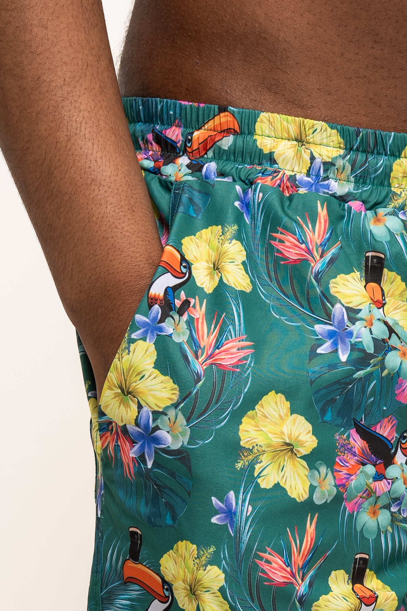 A person wearing Guinness Toucan Hawaiian Green Beach Shorts from Guinness Webstore US, featuring yellow, purple, and orange flowers with toucan illustrations, standing with one hand in the pocket.
