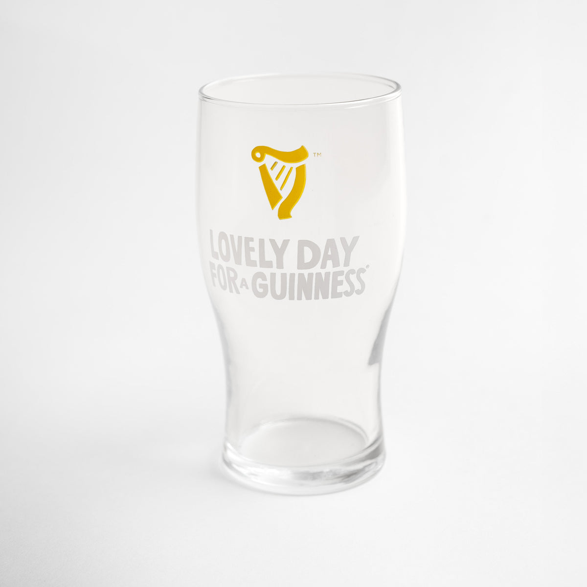 The Guinness Seb Curi Lovely Day Pint Glass from Guinness Webstore US features a yellow harp logo and "Lovely Day for a Guinness" on the front, shown against a plain white background.