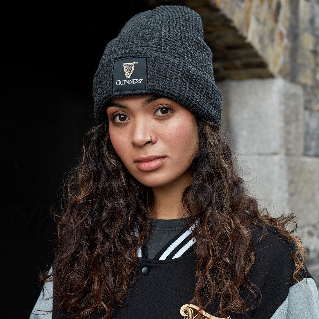 A woman with long curly hair, wearing a black Guinness Thermal Warm Beanie and a black jacket, stands before a stone wall looking at the camera. The unisex Guinness beanie adds a stylish, cozy touch to her outfit.