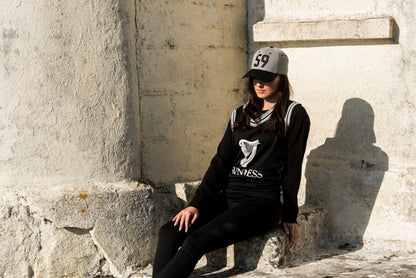 A woman wearing a Guinness Grey 59 Baseball Cap (Adjustable) and black leggings sitting on a stone wall.