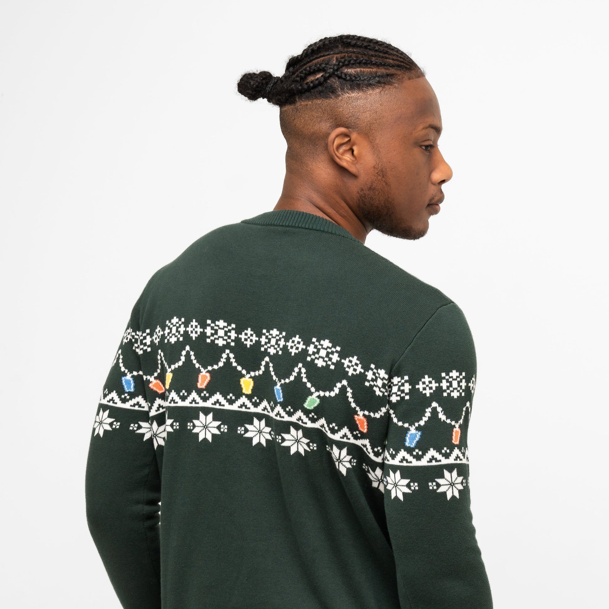 A man with short braids, seen from behind, wears the Guinness Holiday Pint Lights Sweater in green from Guinness Webstore US, featuring festive white snowflakes and colorful holiday light designs.