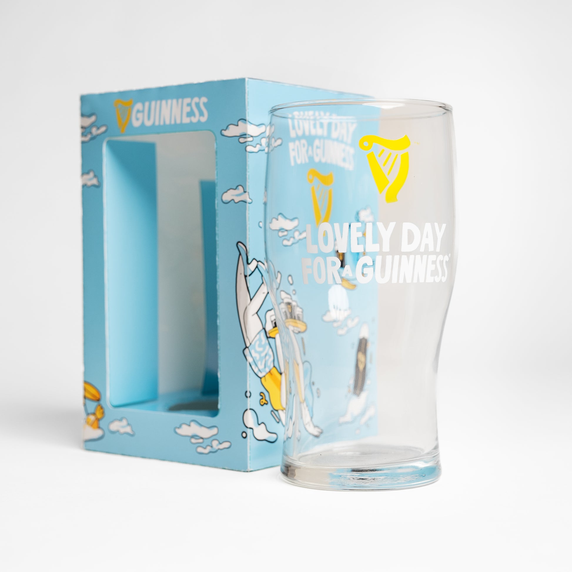 The Guinness Seb Curi Lovely Day Pint Glass from Guinness Webstore US features "Lovely Day for a Guinness" with a yellow harp logo, displayed in front of a blue box with flying toucans and Sebastian Curi's vibrant artwork.