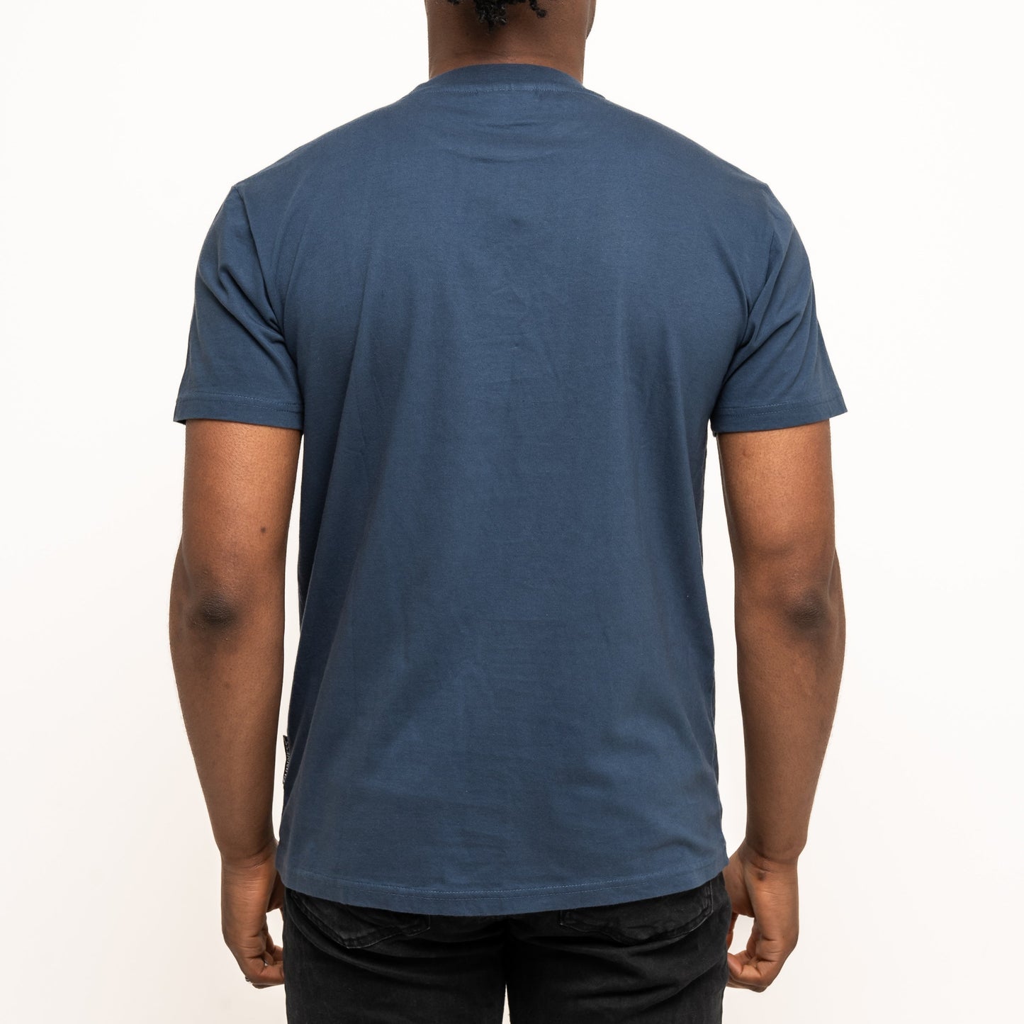 A person with braided hair, wearing black pants and a navy Guinness Transport Collection Tee by Guinness, stands facing away from the camera against a plain white background.