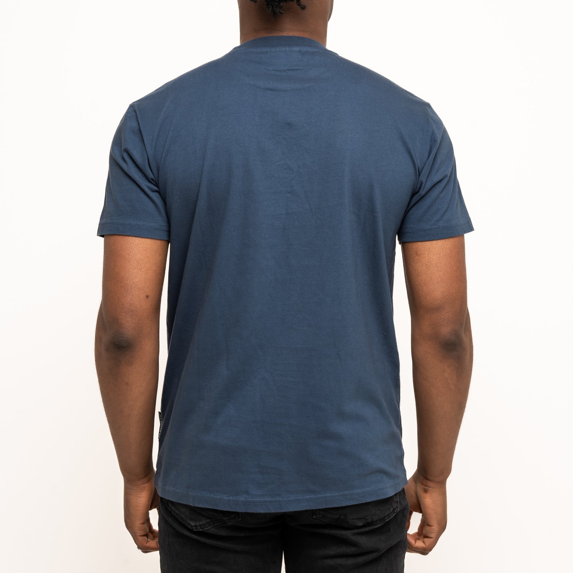 A person with braided hair, wearing black pants and a navy Guinness Transport Collection Tee by Guinness, stands facing away from the camera against a plain white background.
