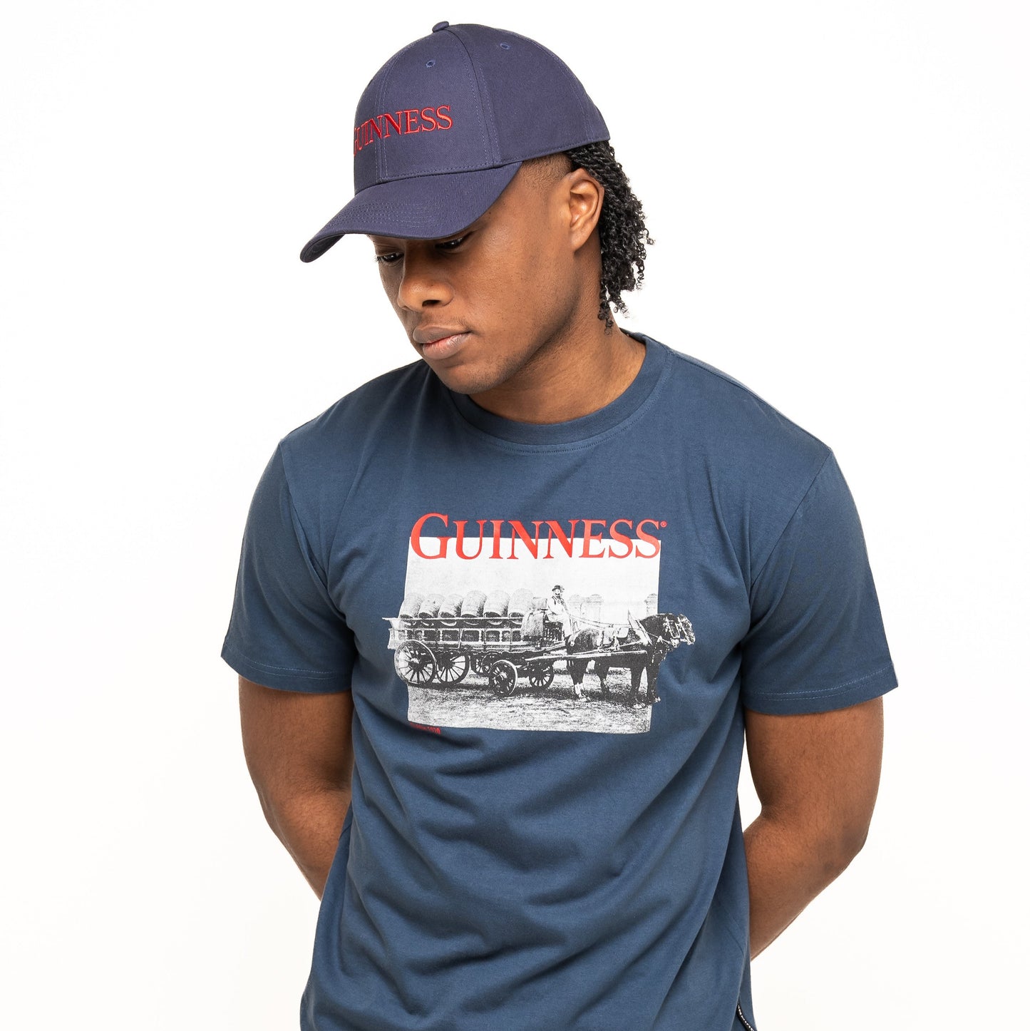 A person wears a navy blue Guinness Transport Collection Baseball Cap from Guinness Webstore US with a matching horse-drawn carriage T-shirt, standing hands behind back against a white background—classic style from St. James’s Gate.