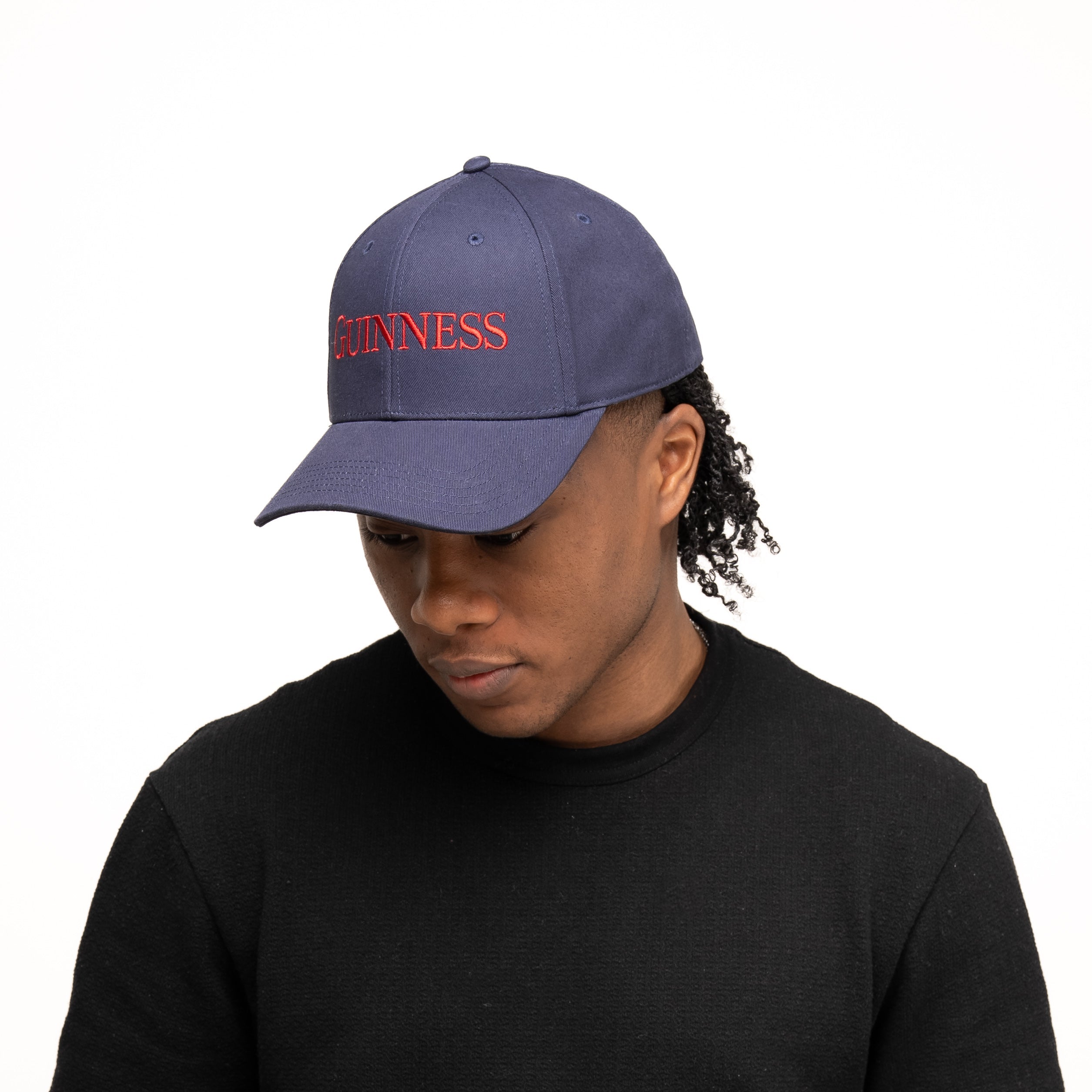 A person wears the Guinness Transport Collection Baseball Cap in blue with red "GUINNESS" lettering from Guinness Webstore US and a black sweater, looking down against a plain white background—evoking classic St. James's Gate style.