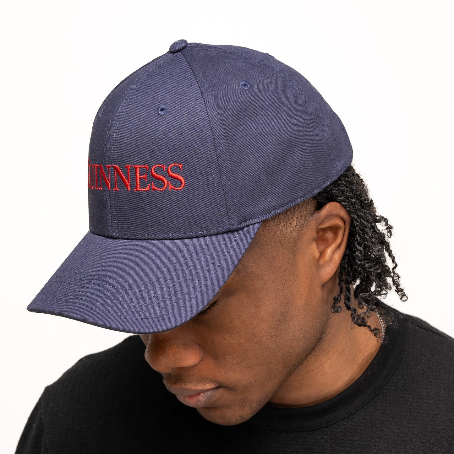 Wearing the Guinness Transport Collection Baseball Cap from Guinness Webstore US, embroidered with "GUINNESS" in red, a person looks downward against a plain backdrop—showcasing classic style inspired by St. James’s Gate.