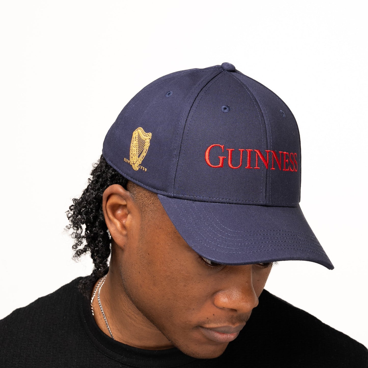 A person wearing the navy blue Guinness Transport Collection Baseball Cap from Guinness Webstore US, featuring "GUINNESS" in red and a gold harp logo, looks downward against a plain background, channeling classic St. James’s Gate style.