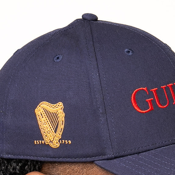 The Guinness Transport Collection Baseball Cap from Guinness Webstore US features navy blue fabric, a gold harp emblem with "ESTD 1759," and partial red "GUI" embroidery on the side, nodding to St. James’s Gate and classic style.