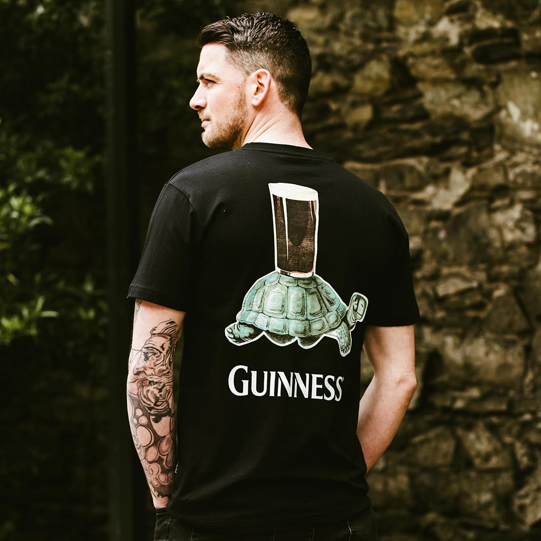 A man with a tattooed arm wears the Guinness Premium Vintage Tortoise Back Graphic Tee—100% cotton, featuring a turtle carrying a pint of Guinness and "Guinness" below—while standing in front of a stone wall.