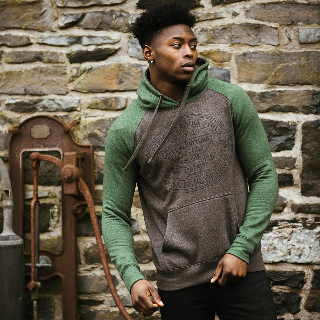 A man wearing a Guinness Grey and Green Hoodie in front of a stone wall while holding a Guinness.