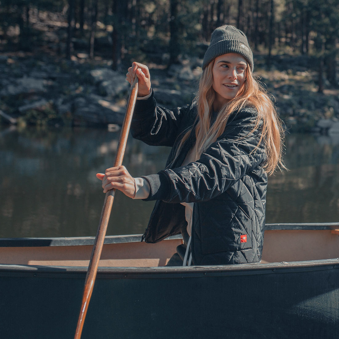 A woman in a Guinness Quilted Utility Zip Up jacket paddles a waterproof black canoe.