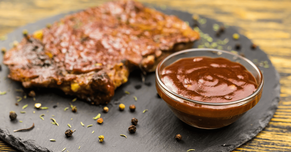A grilled piece of meat with a dark barbecue glaze sits on a black platter next to a small glass bowl filled with Guinness Classic Sauce 295g by Guinness, making it the perfect foodie gift.