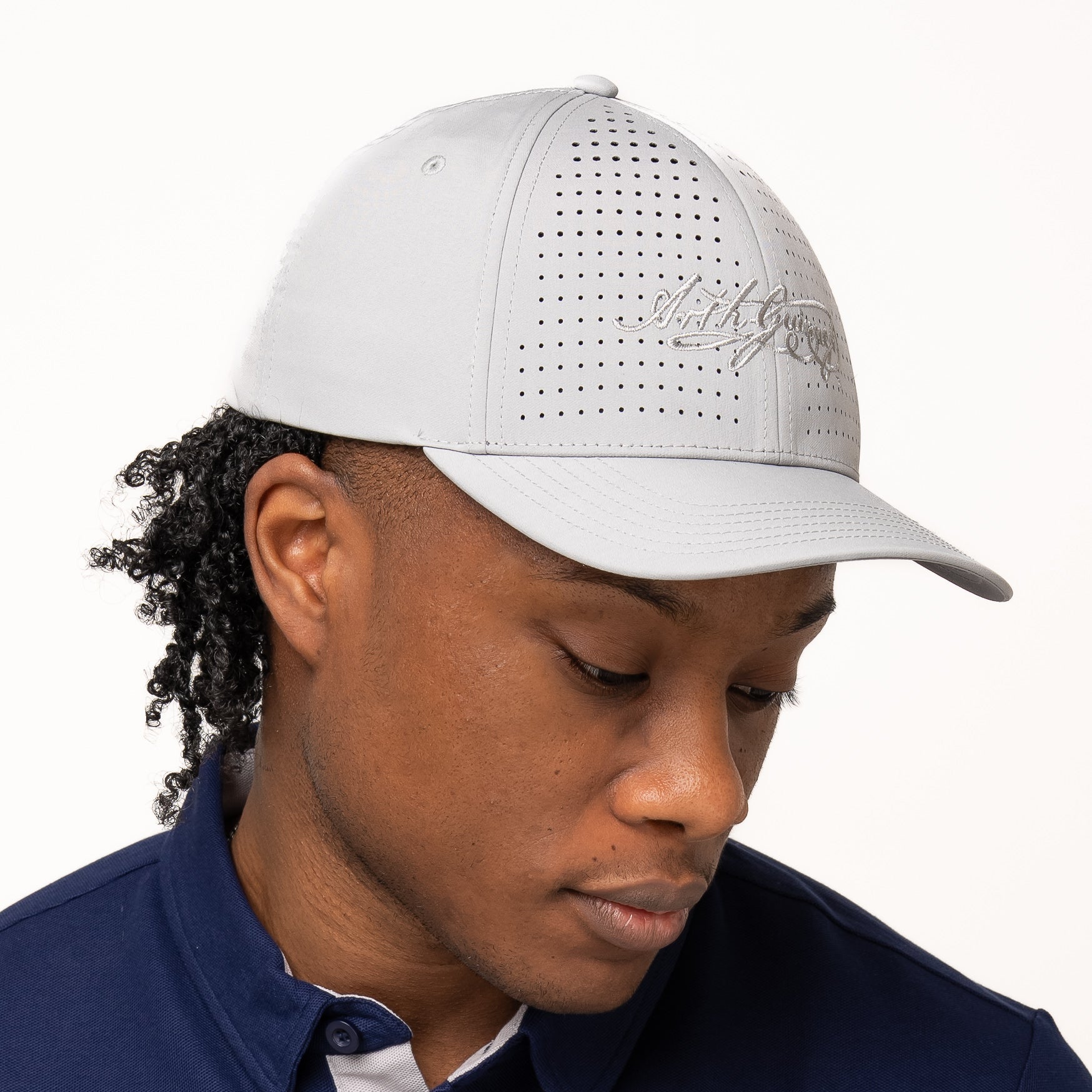 A person wearing a light gray Guinness Performance Signature Cap from Guinness Webstore US, featuring quick-dry technology, and a navy collared shirt with a white harp logo looks down against a plain background.