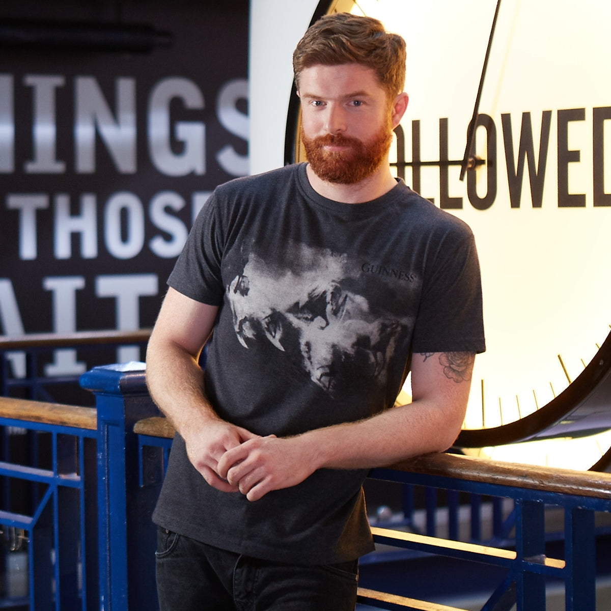 A man leaning against a railing next to a clock, wearing a durable cotton blend fabric Guinness Surf Tee.