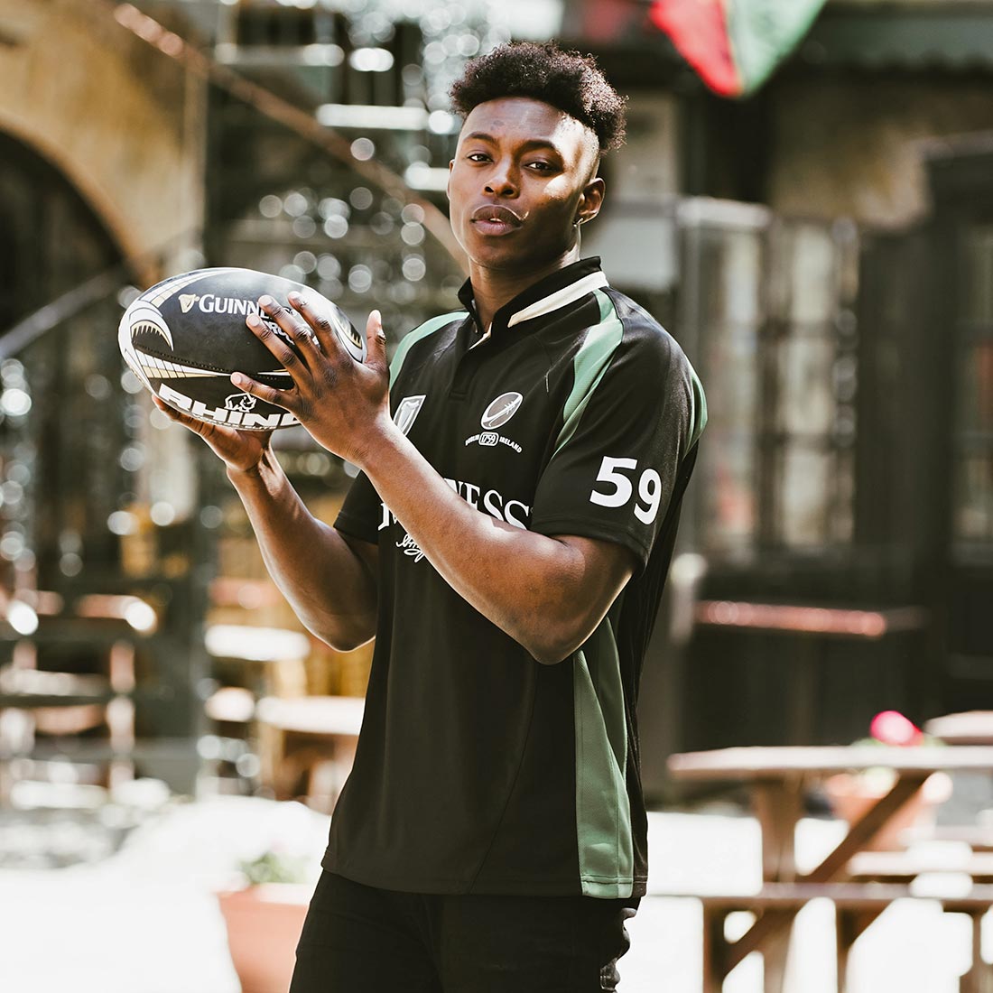 A young man wearing a Guinness Short Sleeve Performance Rugby Jersey, holding a rugby ball in front of a restaurant.