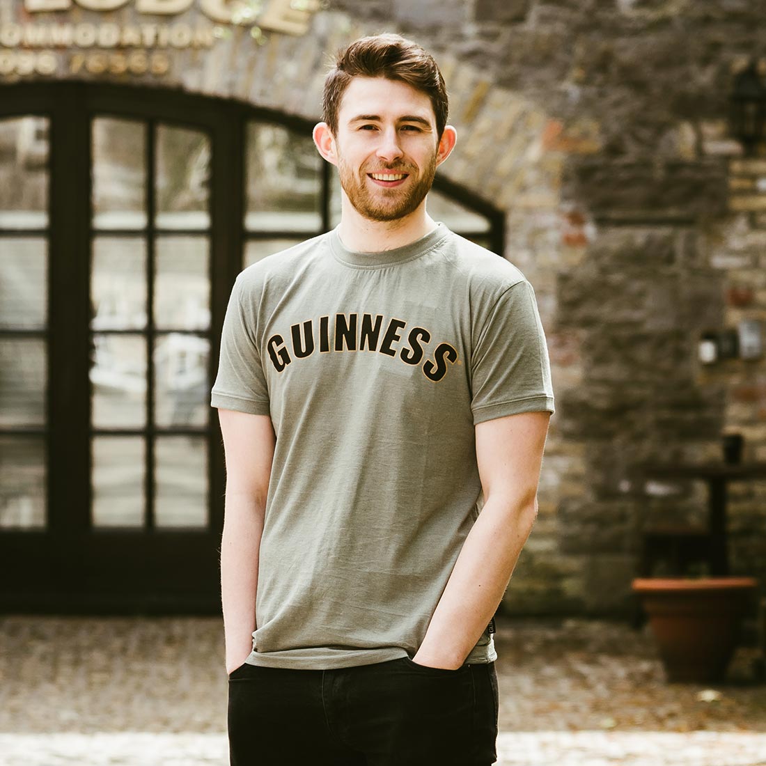 A man wearing a Guinness Green Heathered Bottle Cap Tee in front of a building.