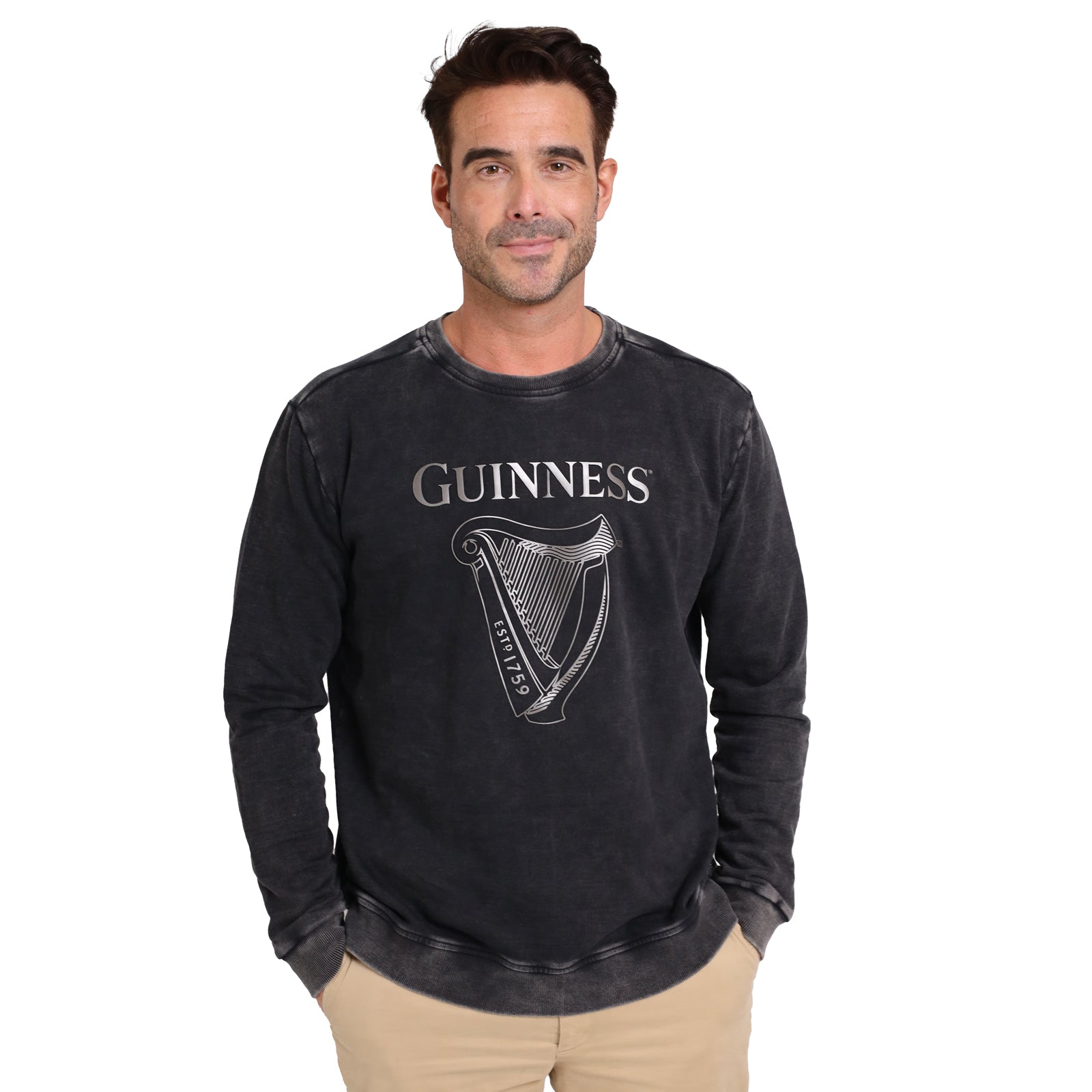 A man stands against a white background wearing the Guinness Embossed Print Trademark Sweater from Guinness Webstore US, featuring a bold harp logo and vintage-style print, paired with beige pants.
