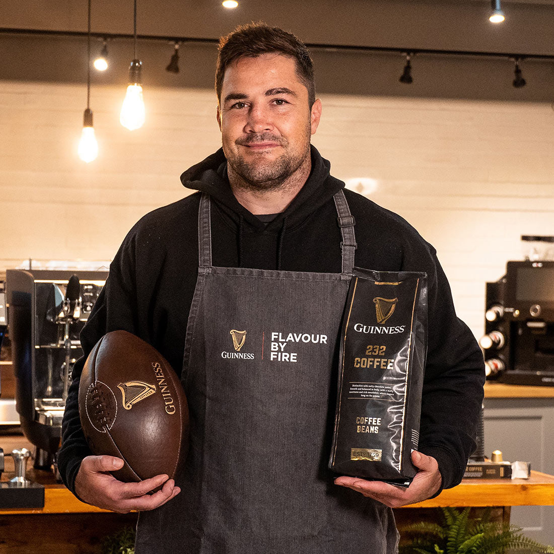 A man in an apron holding a football and a bag of Guinness Coffee Beans 227g.