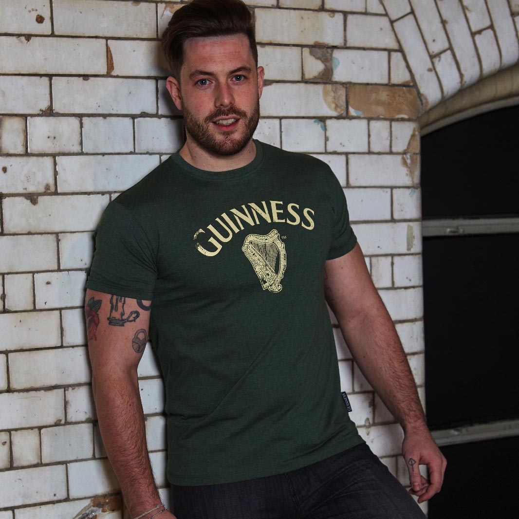A man wearing a Guinness Vintage Harp Tee leaning against a brick wall.