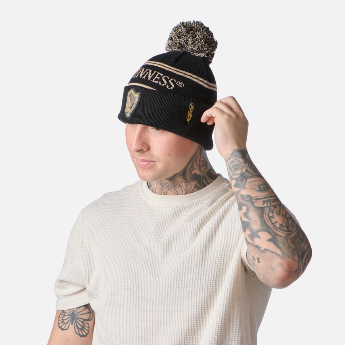 Man in a white shirt adjusts a Guinness Black & Gold Sports Beanie – Vintage Harp Knit; tattoos visible on his arms and neck.