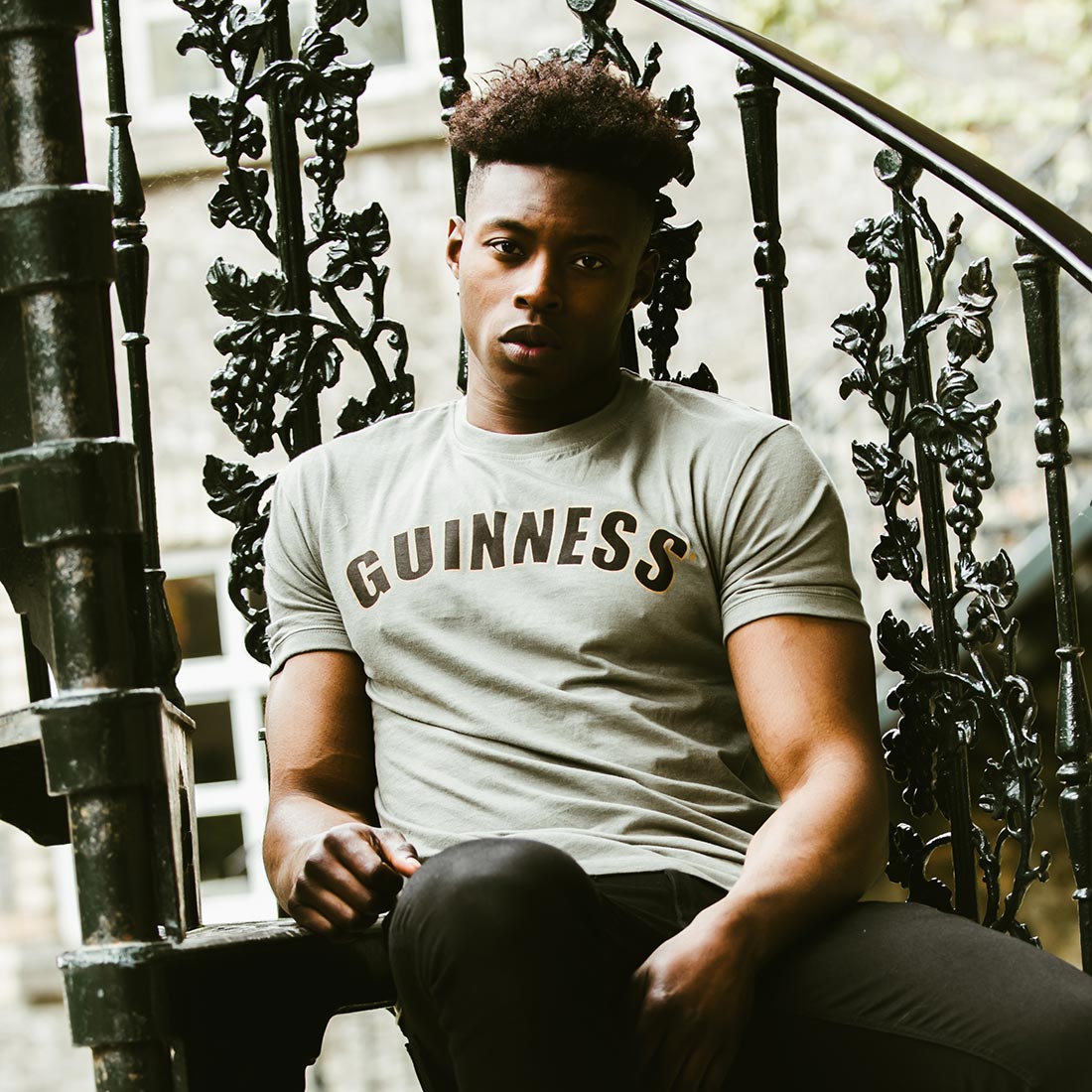 A man wearing a grey Guinness t-shirt, also known as the Green Heathered Bottle Cap Tee, sitting on a staircase.