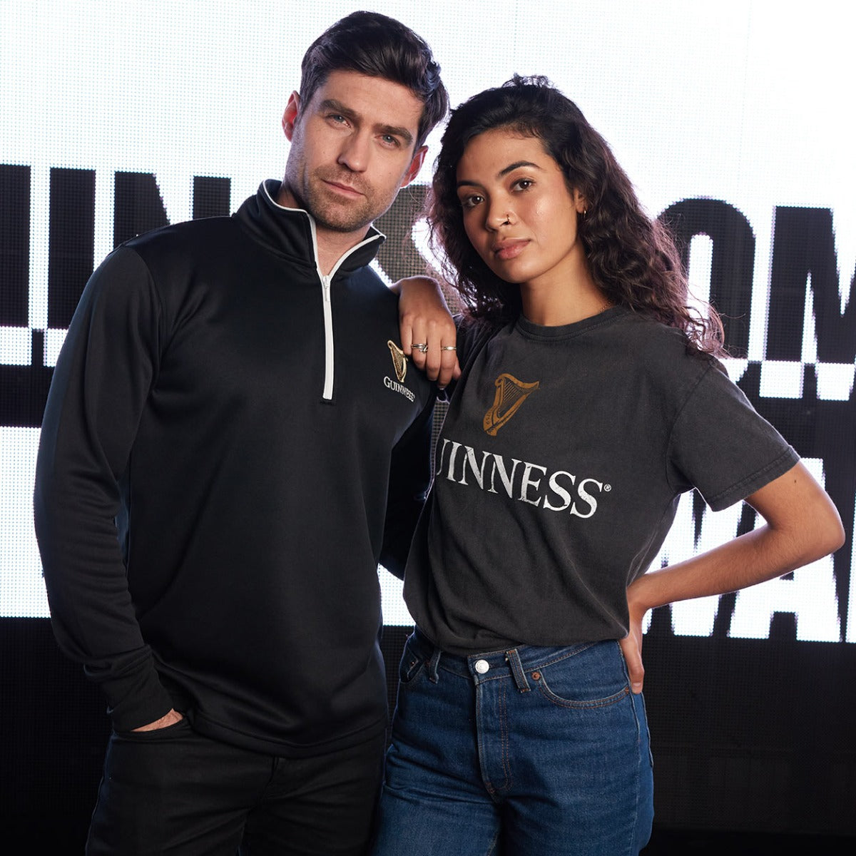 A man and woman, both Guinness fans, posing in front of a Guinness Harp Half Zip Sweater.