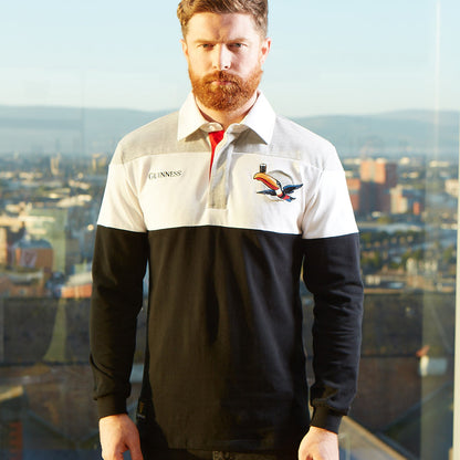 A man with a beard wearing a Guinness Toucan Rugby Jersey standing in front of a city, holding a pint of Guinness while being accompanied by a Toucan.