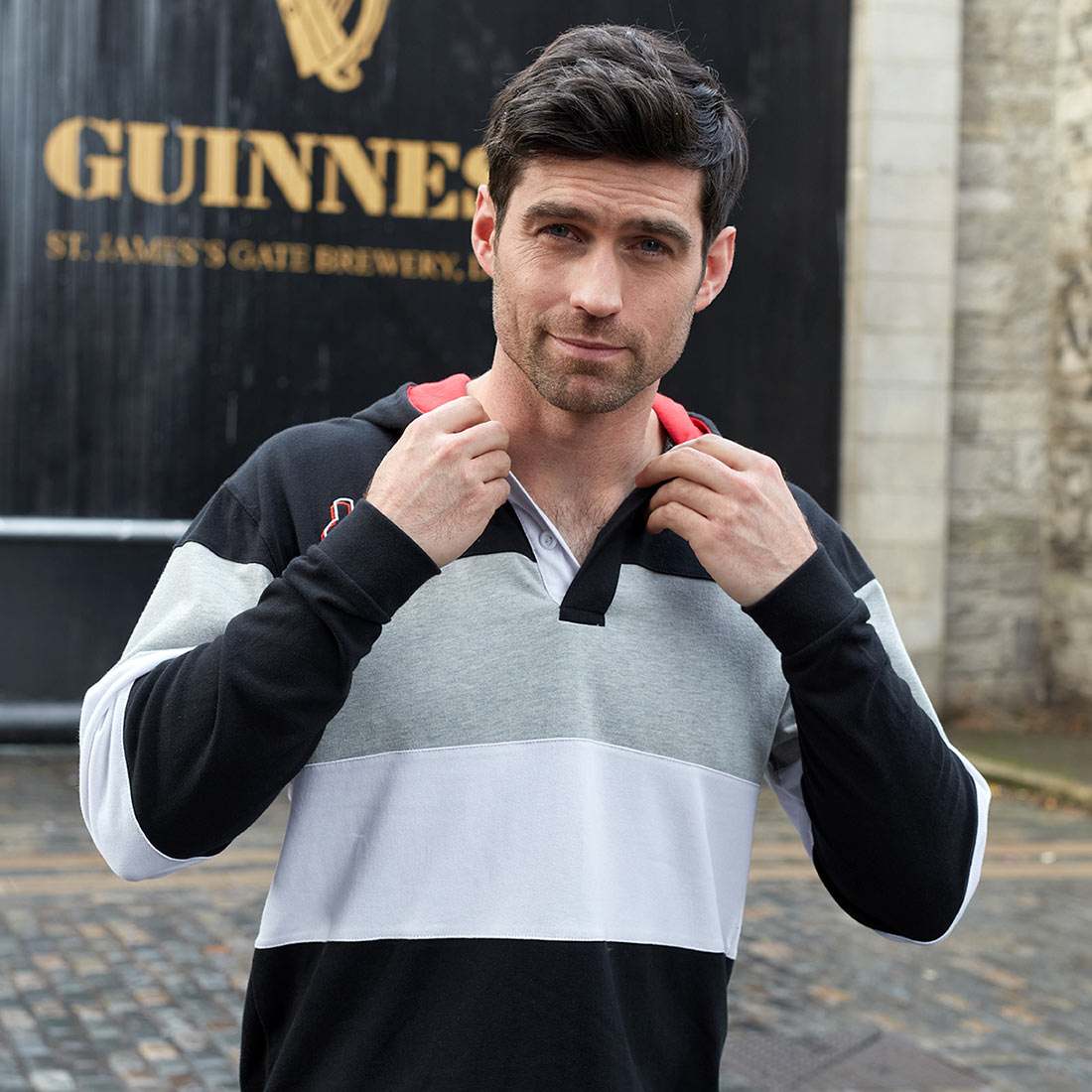 A man sporting a hooded Guinness Black & Red Toucan Hooded Rugby jersey strikes a pose in front of a Guinness sign.